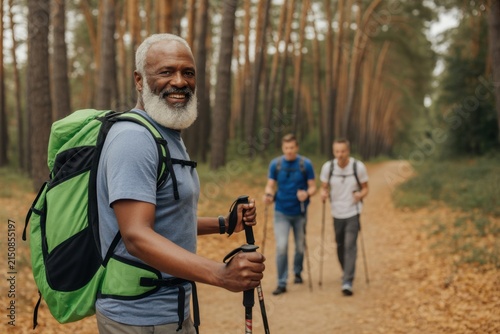 Smiling senior african american man hiking in a pine forest with friends using trekking poles and backpacks for outdoor fitness and active retirement lifestyle activity