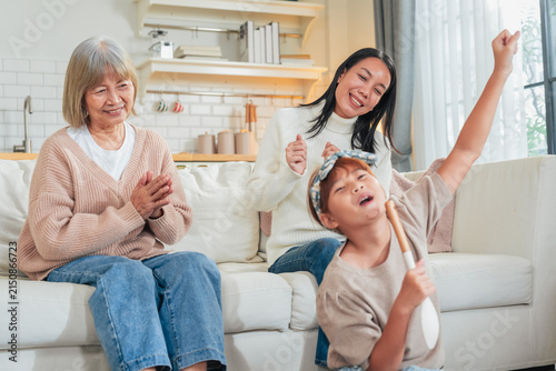 Canvas Print A little girl sings into a wooden spoon microphone while her mother dances and grandmother claps along on the sofa a playful, joyful family moment of impromptu music and shared laughter at home