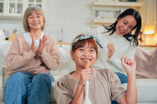 Quadro su tela A little girl sings into a wooden spoon microphone while her mother dances and grandmother claps along on the sofa a playful, joyful family moment of impromptu music and shared laughter at home