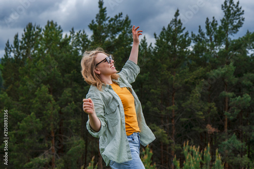 Happy woman in sunglasses standing with raised arm against green pine forest. Cheerful outdoor recreation and active summer lifestyle concept