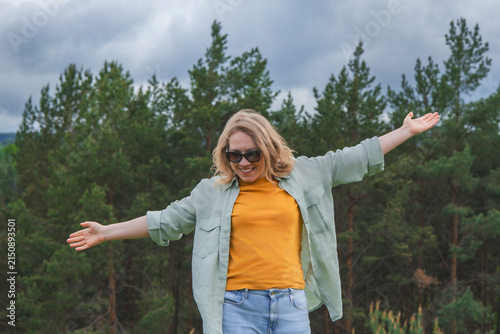 Cheerful woman in sunglasses standing with wide open arms in green forest. Travel freedom inspiration and positive summer emotion concept