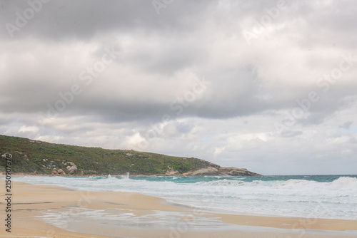 Coastal beach scene featuring a sandy shoreline, rocky headland and gentle ocean waves under cloudy natural light. A clean coastal landscape suitable for travel, nature, tourism and outdoor enviro