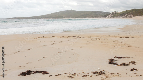 Coastal beach scene featuring a sandy shoreline, rocky headland and gentle ocean waves under cloudy natural light. A clean coastal landscape suitable for travel, nature, tourism and outdoor enviro