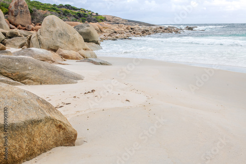 Coastal headland with distinctive red gneiss rocks beside a sandy beach under natural daylight. A rugged shoreline scene showing rock formations, ocean waves, and coastal landscape textures.
