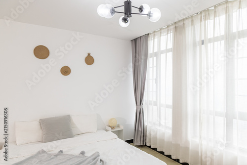Konstfotografi A bright, minimalist residential bedroom interior featuring a bed, decorative wall pieces, sheer curtains, and a modern chandelier
