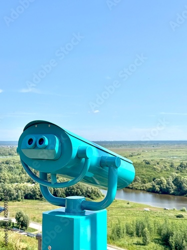 Turquoise coin-operated binocular viewer on a scenic viewpoint overlooking a river and green landscape under a blue sky. Travel and tourism concept.