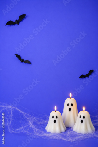Three white ghost candles with flames, black bats and spider web on blue background. Halloween decoration photography with copy space. Halloween holiday concept