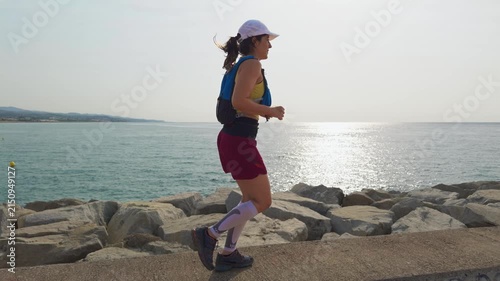 Woman jogs along a rocky coastal path. She wears a white cap, blue vest, red shorts, and black sneakers. The calm ocean stretches wide with sunlight reflecting. Yellow buoys float near the shore