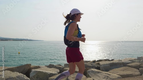 Woman runs along a rocky sea shore. She wears red shorts and a white cap. Her ponytail swings with each step. Sunlight reflects on calm blue water behind her. The scene is bright and open