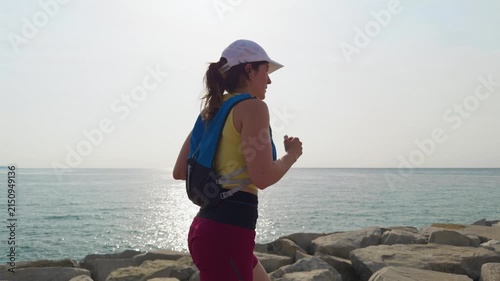Woman runs along rocky sea shore. She wears a white cap and sporty gear. Sunlight glints on the sea water. Her stride shows focus and energy. This scene captures outdoor fitness
