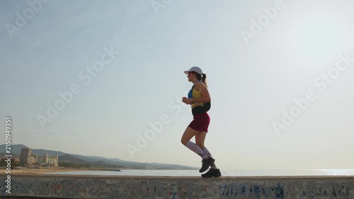 Sunlight glints on the coastal promenade. Runner moves steadily along the path. They wear sportswear and a cap. Their focused steps show determination. Distant buildings and mountains frame the scene