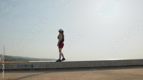 Runner moves steadily on coastal path. Bright sun highlights clear sky. Calm and focused energy. Ideal for fitness or travel videos. Ocean and mountains provide scenic backdrop