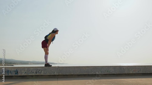Runner stands on a concrete ledge. Calm focus guides her gaze. City and sea meet on the horizon. Her athletic wear suggests steady motion. Mid-stride, her feet strike the pavement
