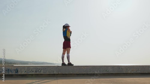 Young woman stands on a concrete pier, facing the vast ocean. She wears red shorts, white socks, and a blue helmet. Arms raised high, eyes closed, serene smile visible. Perfect for travel, adventure