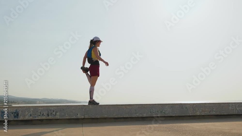 Young woman stretches on a seaside promenade at sunset. Sun glints behind her, casting a warm golden glow. She wears a blue tank top and maroon skirt. Back to camera, facing the calm ocean

