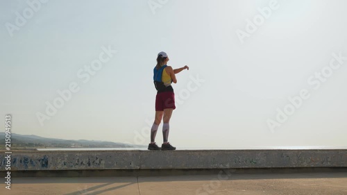 Person stands on concrete seawall, arms raised high. Back to camera, confident pose. Points toward distant ocean. Clear sky, calm sea visible. Happy traveler celebrates new view