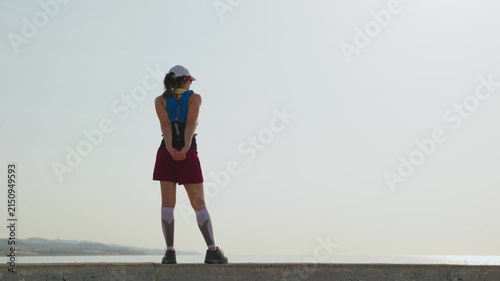 Woman stands alone on a quiet beach. Her blue tank top and maroon skirt are visible. White socks and a cap complete her look. She gazes toward distant hills