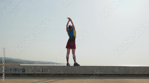 Young woman stretches on a concrete sea ledge. She wears a red skirt and blue top. Her backpack rests on her back. She faces away, arms raised high. Hazy mountains glow behind her