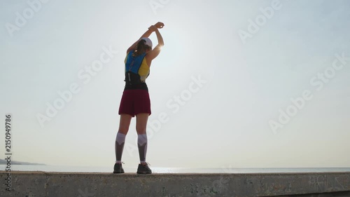 Runner stretches arms high toward sunset. Backpack visible on shoulders. Person appears relaxed and satisfied. Perfect for fitness or wellness content. Calm coastal environment