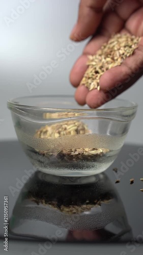 A hand pours golden fennel seeds into a small glass bowl, reflecting on a dark surface, highlighting the process of preparing spices for cooking or traditional remedies