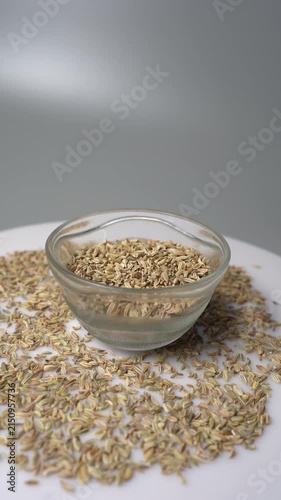 Close-up of Anise Seeds (Pimpinella anisum) in a Glass Bowl on a White Surface with Seeds Scattered Around, Rotating Slowly Against a Gray Background