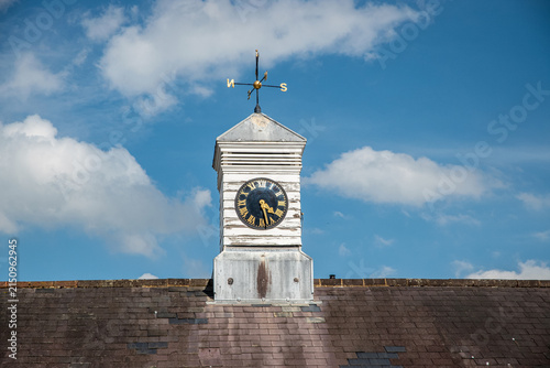 Centered historic clock tower with Roman numeral face and weathervane above a tiled roof in rural England.