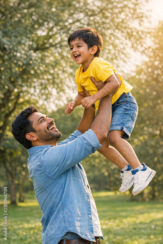 Smiling man holding child aloft on grassy field under sunny sky.