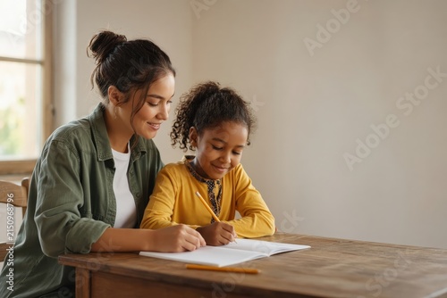 An older girl assists a younger child with writing exercises at a wooden desk.