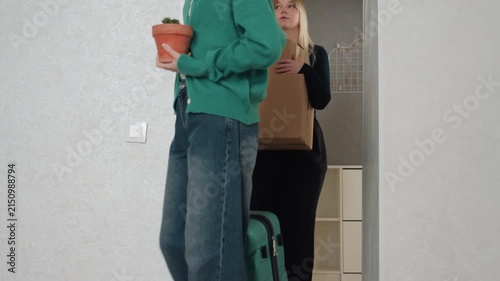 Roommates pause in doorway with boxes, one holds potted plant while other carries stacked cardboard, closeup on feet and hands, soft smiles and tired relief, grey wall and shelving backdrop, scene