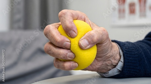 Hand of an elderly person squeezes a yellow stress ball to improve muscle strength during a physical therapy session.
