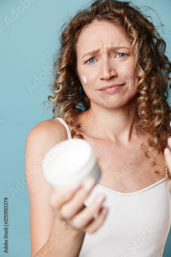 Middle aged woman with natural skin holds a cream jar and looks puzzled at the camera. Studio shot on a blue background. Concept for skincare, beauty routine, or product testing.