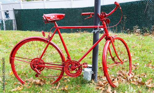 Vintage classic cruiser bicycle painted completely in bright red color locked with a chain to a metal post on a green grass lawn with dry leaves