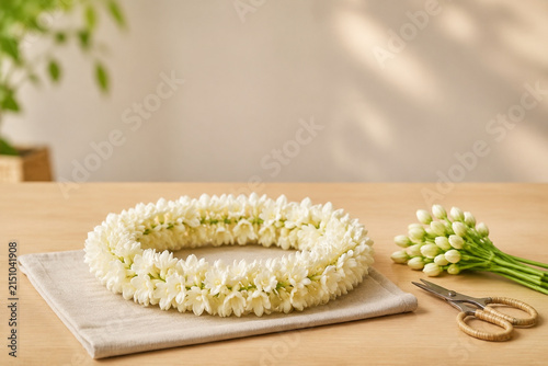 Sampaguita floral garland with buds, craft scissors, and folded linen cloth on wooden table, Filipino floral offering concept