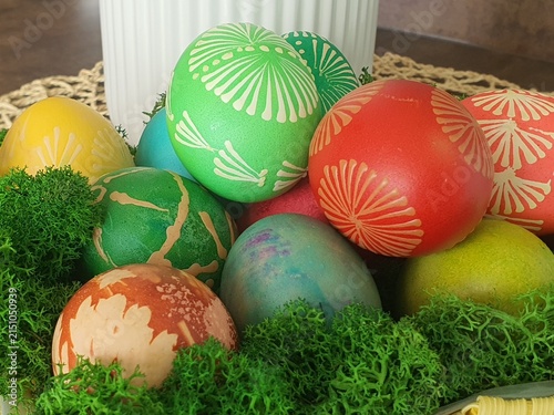 A close-up arrangement of colorful decorated Easter eggs nestled in vibrant green moss. The eggs display various decorative patterns including white floral and radial designs on bright green and red