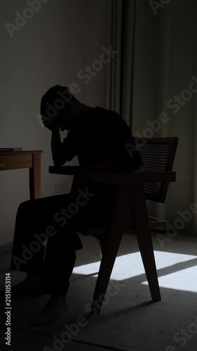 Silhouette of lonely man sitting alone in dark room, lowering his head into his hands in emotional distress, expressing sadness, despair, isolation and inner struggle, vertical