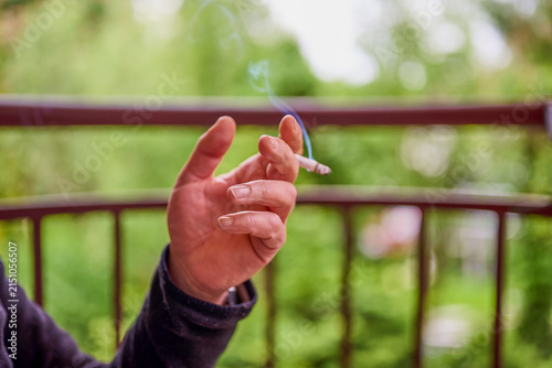 A person is sitting on a balcony and holding a cigarette. Green trees and plants are visible in the background. The sun is shining brightly on this day