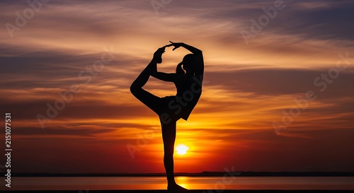 A beautiful silhouette of a woman performing an advanced yoga pose against a vibrant golden sunset reflecting over the calm water at dusk