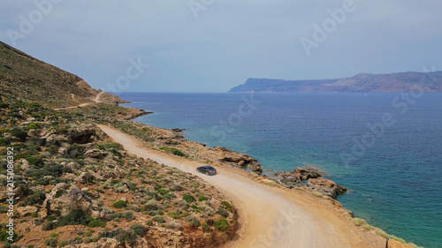 Aerial view of black car driving along coastal dirt road near turquoise sea water during summer road trip in Crete Greece
