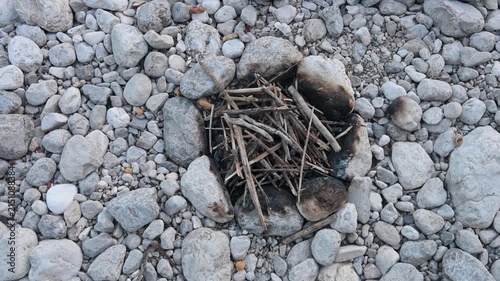 Small rustic campfire pit built with river rocks on a pebble bank. Dry twigs and branches arranged inside a stone fire ring for camping.