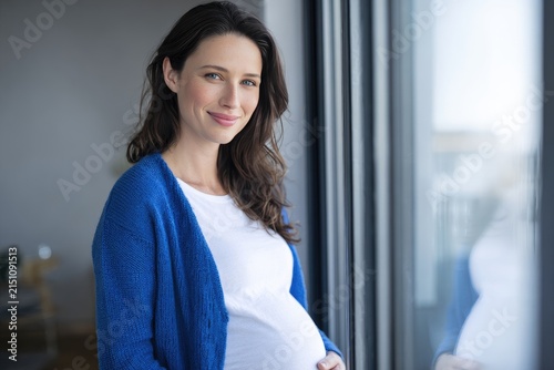 Smiling Pregnant Woman in Blue Cardigan Indoors Near Window