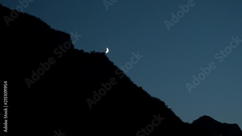 Thin crescent moon shining in a dim twilight sky above a mountainous ridge. Bright crescent moon glowing over a dark mountain slope at dusk.