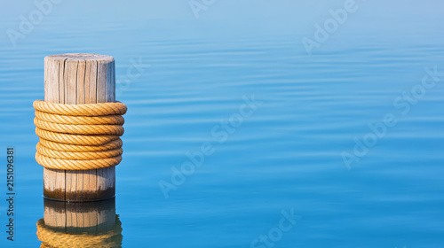 Wooden post with thick nautical rope coiled around it stands calm blue water, creating serene