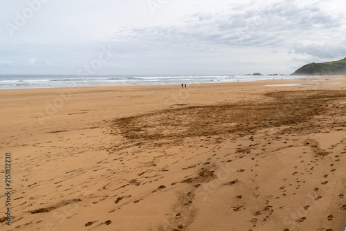 Expansive sandy beach with gentle waves rolling onto the shore under a cloudy sky, featuring distant figures and footprints in the foreground