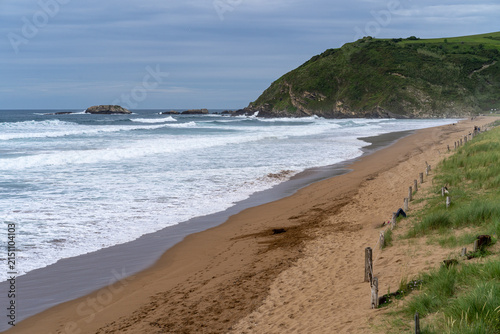 A scenic view of Zarautz beach with waves crashing on the shore and a green hill overlooking the coastline