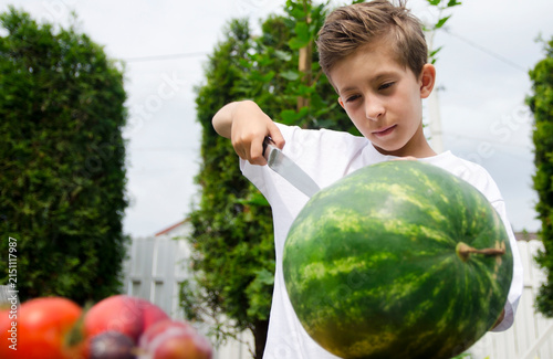 Fotografía Young boy carefully cutting a whole watermelon on a wooden board outdoors