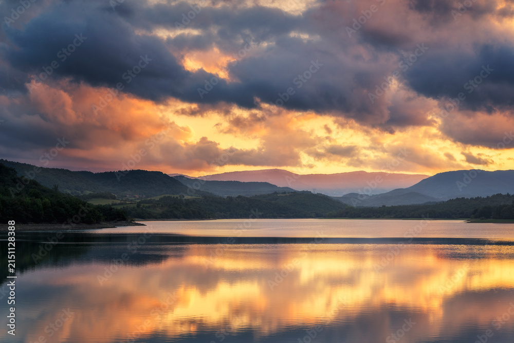 Fototapeta premium Scenic view of Ordunte reservoir at sunset surrounded by mountains under dark dramatic stormy clouds with warm light reflections on calm water