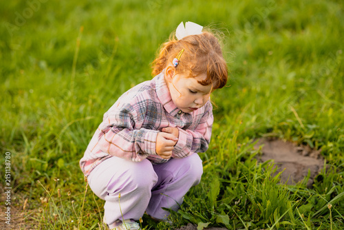 A young girl sits on the grass in a garden. She is looking at the soil with interest and is wearing a pink plaid shirt. It is a sunny afternoon.