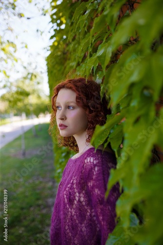 Redhead teenage girl in knitted purple transparent sweater posing at green ivy wall. Clothing and fashion concept