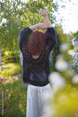 Young girl with red curly hair in black knitted sweater posing at white blossoming bush. Back and side view. Model on spring background