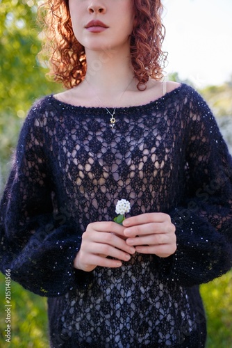 redhead teenager girl in black knitted sweater holding tiny flower. Face partly visible. Model on spring background